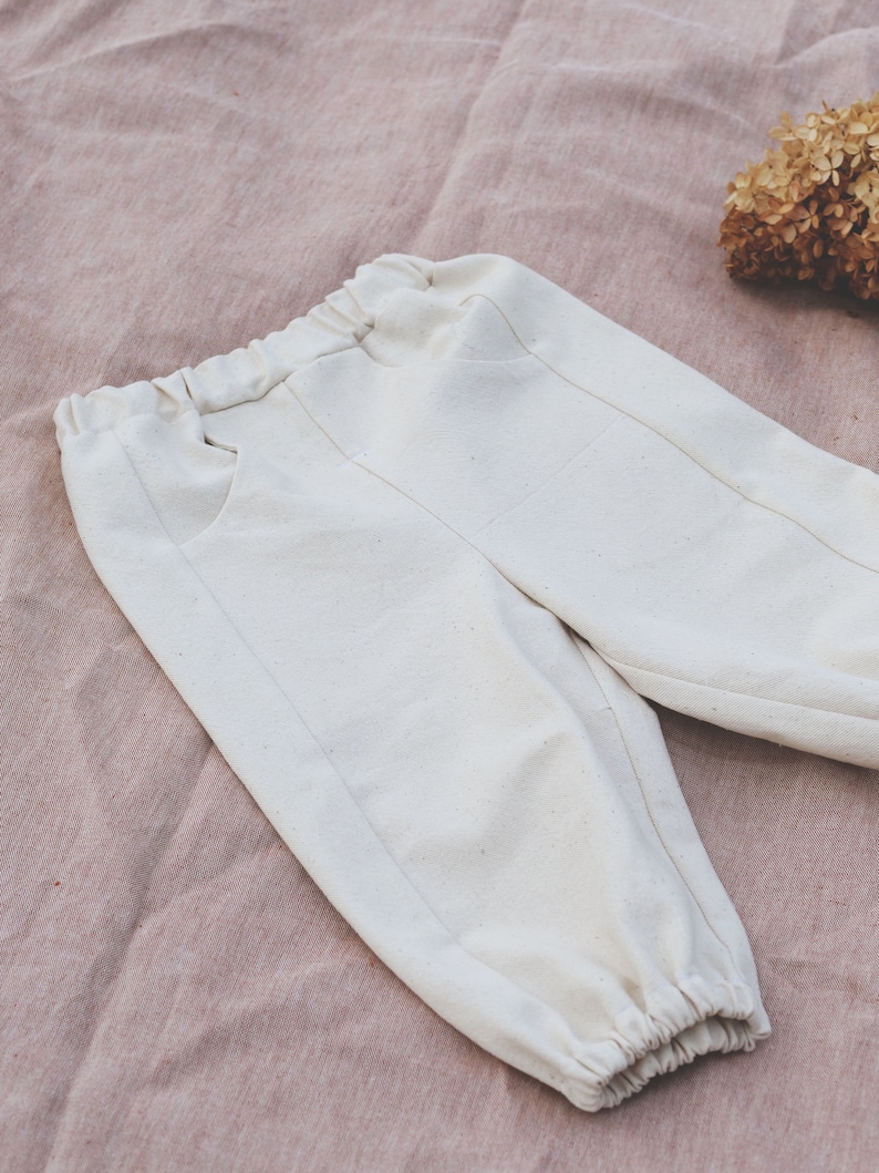 Op de afbeelding: Witte katoenen broek met een trekkoord in de taille en elastische boorden. De broek heeft twee zakken aan de voorkant.