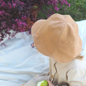 May include: A child wearing a brown linen sun hat and a beige linen dress holds a green apple. The child is sitting on a white blanket.