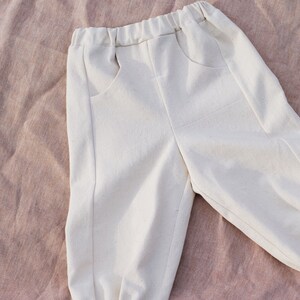 Op de afbeelding: Witte katoenen broek met een trekkoord in de taille en elastische boorden. De broek heeft twee zakken aan de voorkant.