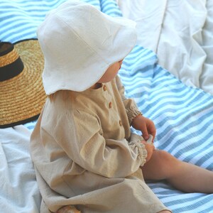 May include: A child wearing a white linen sun hat and a beige linen dress sits on a striped blanket. A loaf of bread is on the blanket next to the child.