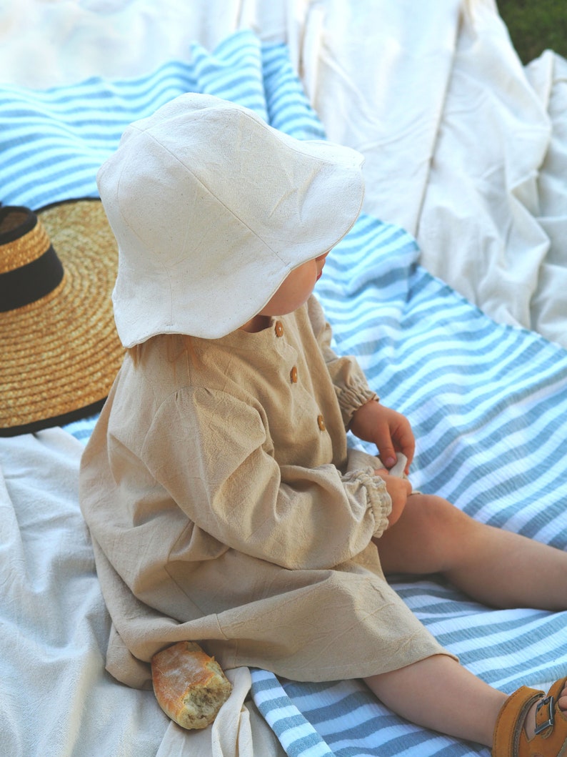May include: A child wearing a white linen sun hat and a beige linen dress sits on a striped blanket. A loaf of bread is on the blanket next to the child.