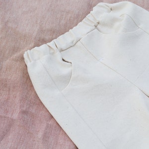 Op de afbeelding: Een witte katoenen broek met een trekkoord in de taille en zijzakken. De broek is gevouwen en ligt op een roze stoffen achtergrond.