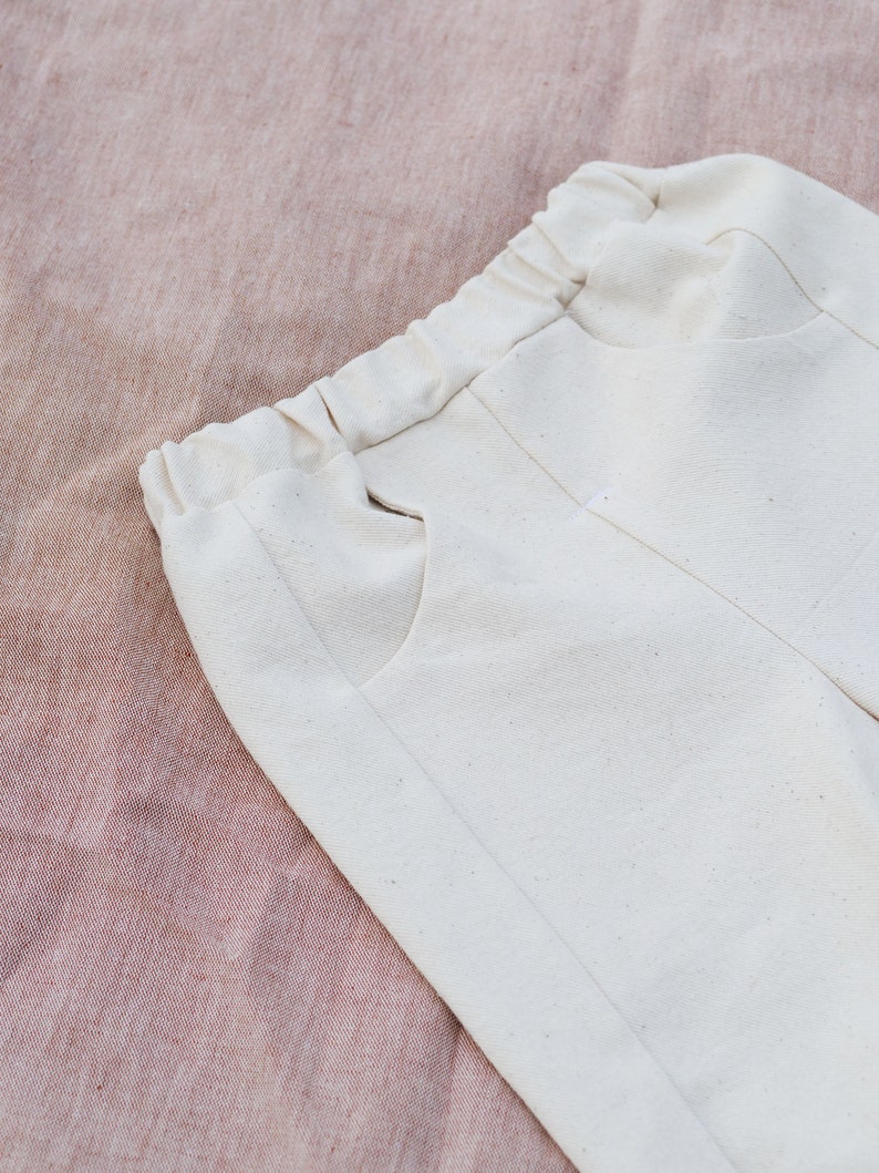 Op de afbeelding: Een witte katoenen broek met een trekkoord in de taille en zijzakken. De broek is gevouwen en ligt op een roze stoffen achtergrond.