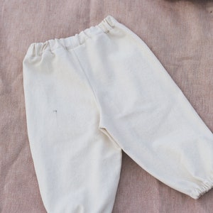 Op de afbeelding: Een witte katoenen broek met een trekkoord in de taille en elastische boorden. De broek ligt plat op een roze stoffen achtergrond.