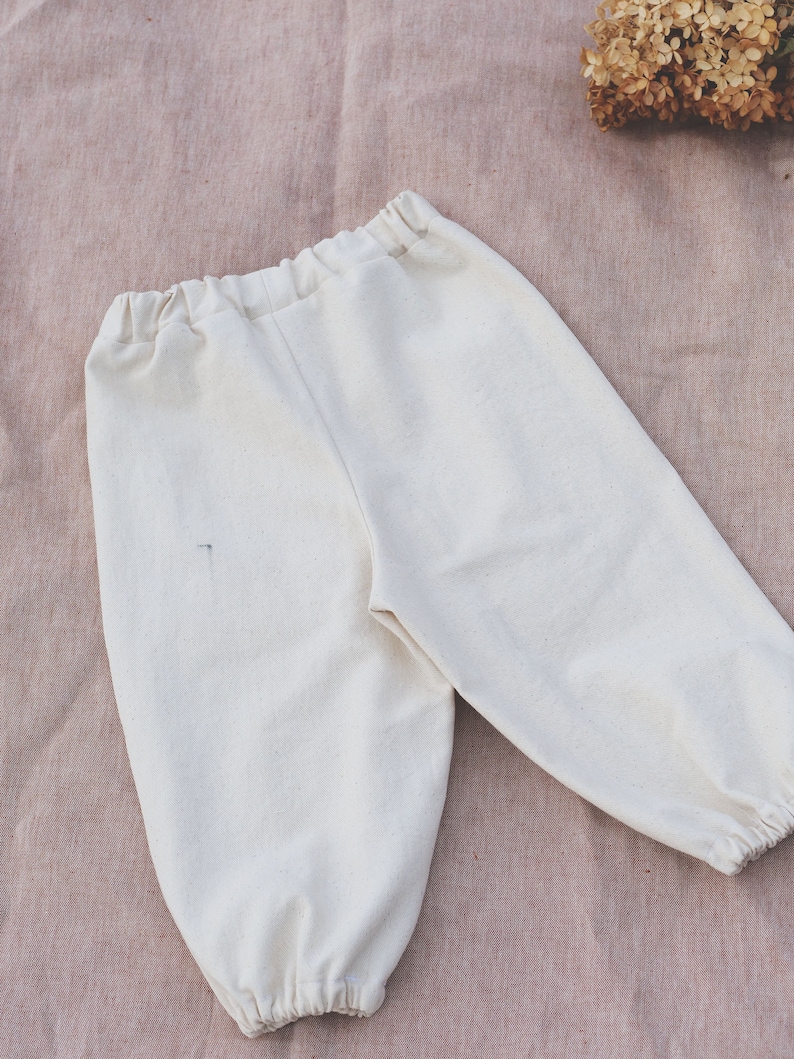 Op de afbeelding: Een witte katoenen broek met een trekkoord in de taille en elastische boorden. De broek ligt plat op een roze stoffen achtergrond.