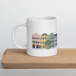 May include: White ceramic mug featuring a colorful illustration of historic buildings in shades of pink, yellow, green, and gray. The buildings have black shutters and are lined up in a row. A tree is between the yellow and green buildings.