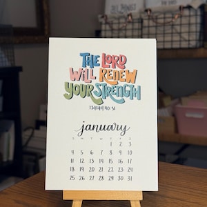 May include: A small, white desk calendar for January, displayed on a wooden easel. The calendar features colorful text that reads "THE LORD WILL RENEW YOUR STRENGTH" and the scripture reference Isaiah 40:31. The calendar grid is in black.