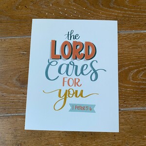 Peut inclure: Une impression de couleur bleu clair avec le texte "the LORD cares for you" en lettres stylisées colorées. Le texte "1 Peter 5:6" est également inclus dans une police plus petite.