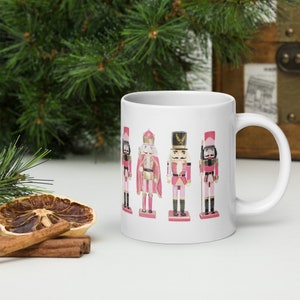 Könnte beinhalten: Weiße Keramiktasse mit einem rosa und goldenen Design mit fünf Nussknackerfiguren. Die Nussknacker tragen rote und goldene Uniformen mit rosa Akzenten.