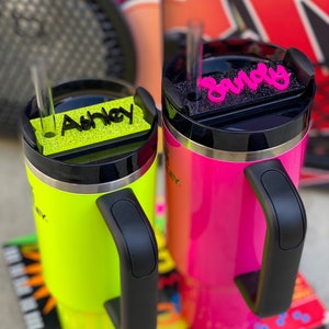 May include: Two insulated tumblers with black handles and lids. The left tumbler is neon yellow with a neon yellow glitter name tag reading Ashley. The right tumbler is neon pink with a black glitter name tag reading Sandy. Both have clear straws.
