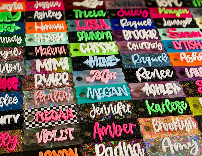 May include: A collection of rectangular acrylic keychains featuring various names in raised, colorful 3D lettering. The base plates display diverse finishes including glitter, solid colors, and checkered patterns, each with a circular hole for attachment.