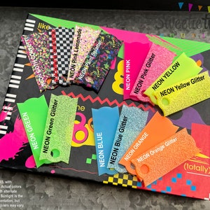 May include: A collection of rectangular acrylic color swatches with rounded edges and a hole at the bottom. The swatches feature various neon colors, glitter finishes, and patterns including checkered, confetti, and paint splatter with printed labels.