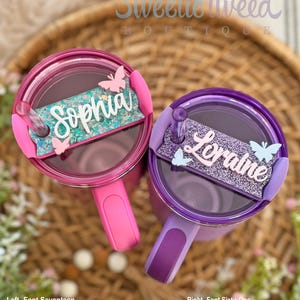 May include: Two personalized tumblers with butterfly accents. The pink tumbler has the name "Sophia" on it and the purple tumbler has the name "Lorraine" on it. Both tumblers have a straw and a handle.