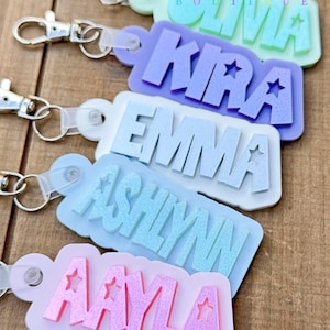 May include: Five pastel-colored keychains with names spelled out in bold letters and silver star accents. The names are Olivia, Kira, Emma, Ashlynn, and Aayla. The keychains are attached to silver split rings.