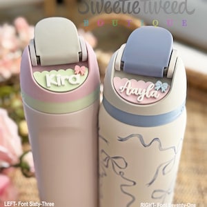 May include: Two insulated water bottles with flip-top lids. The left bottle is light pink with a pastel green name plate reading Kira and a small pink bow. The right bottle is white with blue ribbon patterns, a pastel pink name plate reading Aayla and a blue bow.