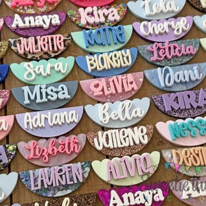 May include: A collection of semi-circular name badges featuring various names in raised 3D lettering. The badges display diverse finishes including glitter, solid colors, and marbled patterns, with scalloped edges along the top and bottom.