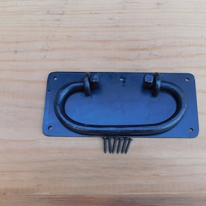 May include: Black metal door pull with a rounded handle and a rectangular base. The pull is attached to a wooden surface with screws.