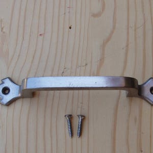 May include: A silver-toned metal door handle with a decorative design on each end. The handle is mounted on a wooden surface and includes two screws.