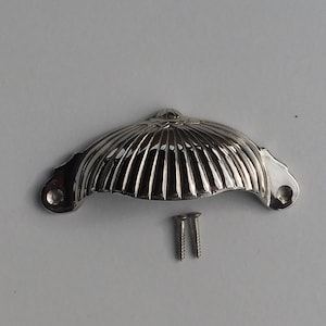 Puede incluir: Un tirador de armario de forma de concha, de color plateado, con un diseño acanalado. Se incluyen dos tornillos plateados.