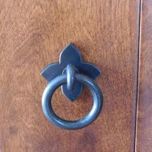 May include: Black metal door handle with a decorative ring pull and a flower-shaped design.