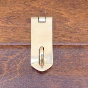 Puede incluir: Un pestillo de metal dorado con un anillo plateado. El pestillo está unido a una superficie de madera.