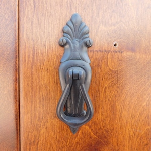 以下が含まれることがあります： 木製のドアに取り付けられた、黒い装飾的なドアノッカー。 ドアは、暖かく赤褐色の仕上げが施された垂直の木製パネルでできています。 ノッカーは、葉のような装飾的な上部を持っています。