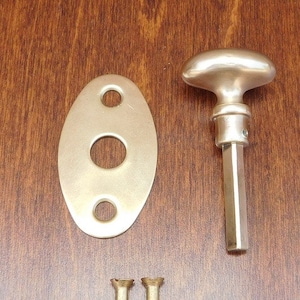May include: A silver door knob with a rounded oval shape and a straight shaft. The knob comes with a matching oval-shaped backplate with three holes and two gold screws.
