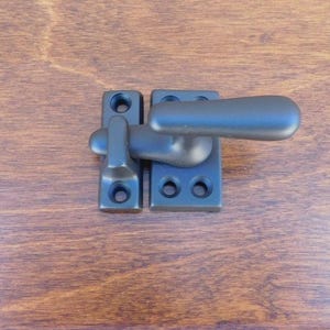 Puede incluir: Pestillo de ventana negro con una manija de palanca. El pestillo está montado en una superficie de madera.