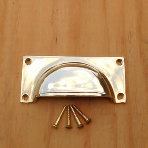 May include: A polished brass drawer pull with a half-moon design, featuring a rectangular backplate with four screw holes. Includes four brass screws. The hardware is set against a light brown wooden surface.
