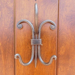 May include: A decorative metal hook with a double-loop design, featuring a twisted, ornate shape. The hook is attached to a wooden door.