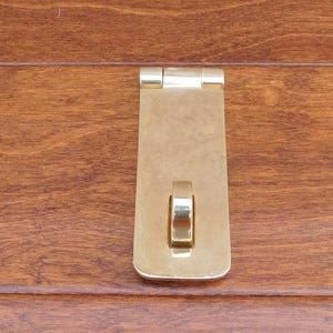 Könnte beinhalten: Ein goldfarbener Türverschluss aus Metall mit abgerundeter, rechteckiger Form. Der Verschluss hat einen kleinen, runden Metallring an der Vorderseite.