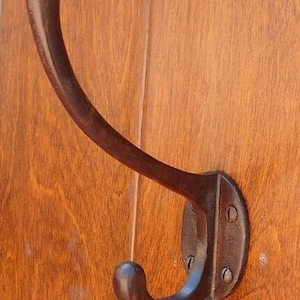 Puede incluir: Un gancho de metal marrón oscuro con un diseño curvo. El gancho está montado en una superficie de madera.