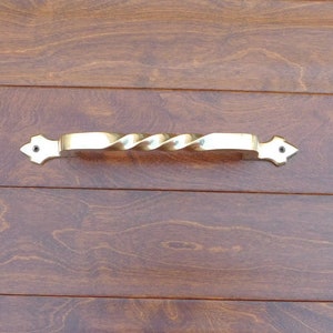 May include: A gold-colored, twisted metal handle with decorative end pieces is mounted on a dark brown wooden surface. The handle's design features a central, spiraled grip and is likely for a cabinet or drawer.