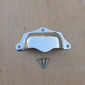 May include: A shiny chrome-finished cup pull with three screws, set against a light wood grain background. The hardware has a curved design with mounting holes on each end, suitable for cabinets or drawers.