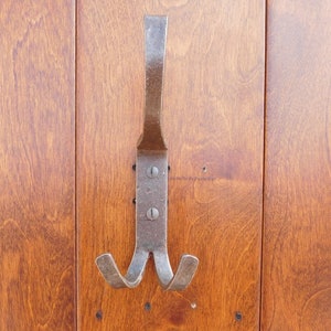 Op de afbeelding: Een enkele, rustieke metalen haak met een dubbelpuntig uiteinde, gemonteerd op een houten oppervlak.