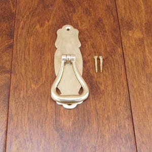 May include: A silver door knocker with a decorative design. The knocker is attached to a wooden door with two screws.