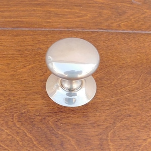 May include: A shiny, silver-colored cabinet knob with a rounded top and a circular base. The knob is set against a warm, brown wooden surface, creating a contrast in textures and colors. This hardware is suitable for furniture restoration or new builds.