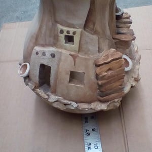 May include: A ceramic vase shaped like a pueblo dwelling with brown, tan, and white paint. The vase has a wide opening and a rounded bottom.