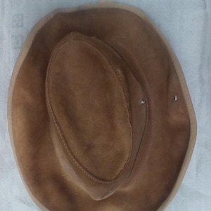 Puede incluir: Sombrero de vaquero de ante marrón con corona redondeada y ala ancha. El sombrero tiene un ribete marrón claro alrededor del ala y está asegurado con dos pequeños remaches. Un clásico estilo western.