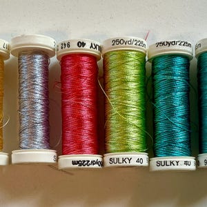 May include: Six spools of Sulky 40 weight embroidery thread arranged in a row. The colors from left to right are yellow, light purple, red, lime green, teal, and dark green. Each spool features white plastic ends with printed text.