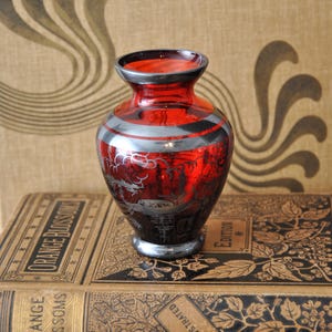 以下が含まれることがあります： 赤色のガラス花瓶、シルバーのアクセントと黒い縁取り。花瓶は丸みを帯びた形で、エッチングされたシルバーのデザインで装飾されています。 金色の文字が入ったヴィンテージ本の山の上に置かれています。