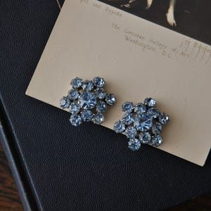 May include: A pair of vintage, light blue rhinestone cluster earrings. The earrings are flower-shaped, with a large central stone surrounded by smaller stones. The earrings rest on a vintage postcard with the text "Cupid and Psyche".