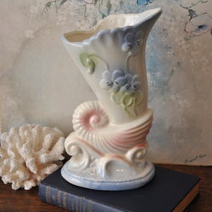 May include: A vintage ceramic vase in a seashell design. The vase is primarily cream-colored with pink, blue, and green accents. It features a flared top and a base shaped like a seashell. The vase is approximately 8 inches tall.