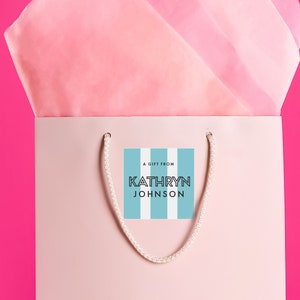 Puede incluir: Una bolsa de regalo rosa con un asa de cuerda blanca. Una etiqueta de regalo a rayas azules y blancas est&aacute; unida a la bolsa con el texto "Un regalo de Kathryn Johnson".