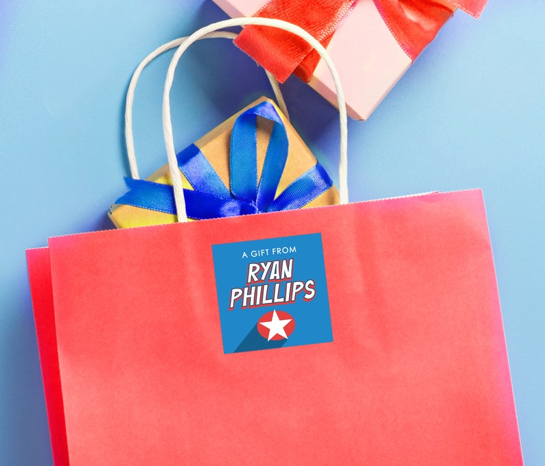 Puede incluir: Una bolsa de regalo roja con asas blancas y una etiqueta de regalo azul y roja que dice "Un regalo de Ryan Phillips".