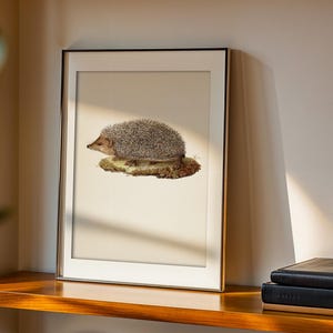 Pode incluir: Impressão artística emoldurada com uma ilustração detalhada de um ouriço. O ouriço, com seu pelo espinhoso cinza e marrom, está empoleirado em um pedaço de musgo. A obra de arte é definida em um fundo creme, emoldurada em preto e dourado.