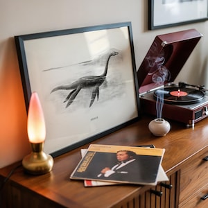 Pode incluir: Uma ilustra&ccedil;&atilde;o em preto e branco emoldurada do Monstro do Lago Ness, rotulada como "Nessie", est&aacute; em um arm&aacute;rio de madeira. Um toca-discos vintage com a parte superior vermelha est&aacute; aberto, tocando um disco. Uma pequena l&acirc;mpada com base dourada e abajur rosa e branco est&aacute; acesa.