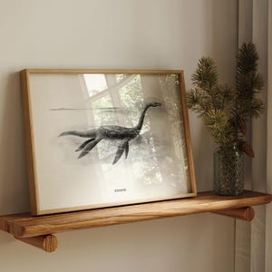 Pode incluir: Uma impress&atilde;o emoldurada com uma ilustra&ccedil;&atilde;o a preto e branco do Monstro do Lago Ness, com a palavra "Nessie" impressa na parte inferior. A obra de arte &eacute; exibida numa prateleira de madeira, ao lado de um vaso com ramos de pinheiro.