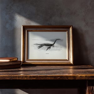 Pode incluir: Ilustra&ccedil;&atilde;o a preto e branco emoldurada de um plesiossauro, um r&eacute;ptil marinho pr&eacute;-hist&oacute;rico, numa moldura dourada. A obra de arte &eacute; exibida numa superf&iacute;cie de madeira escura, com livros &agrave; esquerda. A imagem evoca uma est&eacute;tica vintage.