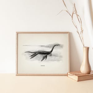 Pode incluir: Ilustra&ccedil;&atilde;o a preto e branco emoldurada do Monstro do Lago Ness, ou Nessie, nadando num lago. A obra de arte est&aacute; numa moldura de madeira e apresenta a palavra "NESSIE" na parte inferior. A imagem tem uma est&eacute;tica vintage.
